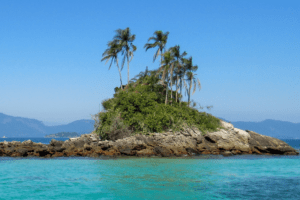 Ilhota rochosa coberta por vegetação e palmeiras, cercada por mar azul-turquesa, com montanhas ao fundo e céu azul limpo.
