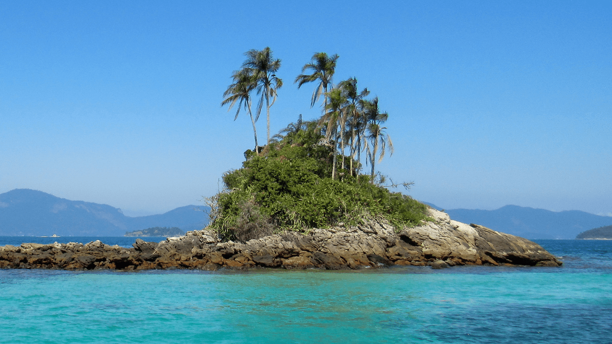 Ilhota rochosa coberta por vegetação e palmeiras, cercada por mar azul-turquesa, com montanhas ao fundo e céu azul limpo.