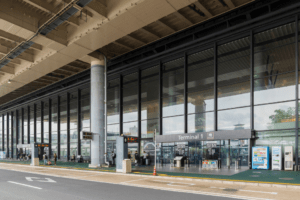 Fachada externa do Terminal 1 de um aeroporto, com área de embarque e desembarque, fachada de vidro e passageiros circulando na entrada