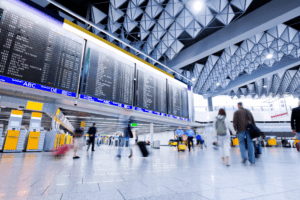 Passageiros circulam no saguão de um aeroporto com painéis de voos ao fundo