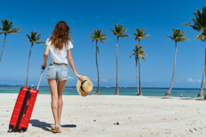 Pessoa caminhando na praia com mala de viagem, segurando um chapéu, com coqueiros ao fundo.