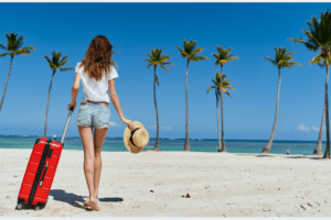 Pessoa caminhando na praia com mala de viagem, segurando um chapéu, com coqueiros ao fundo.