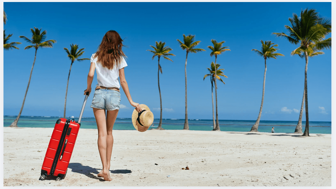 Pessoa caminhando na praia com mala de viagem, segurando um chapéu, com coqueiros ao fundo.