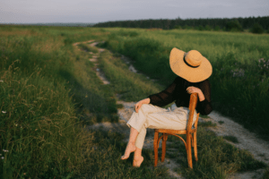 Pessoa sentada em uma cadeira no meio de um campo com vegetação alta, usando chapéu e voltada para uma estrada de terra