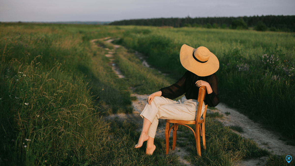Pessoa sentada em uma cadeira no meio de um campo com vegetação alta, usando chapéu e voltada para uma estrada de terra