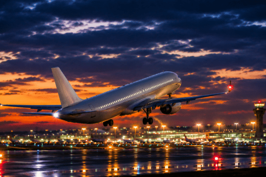Avião comercial decolando ao entardecer em aeroporto iluminado, com pista molhada refletindo as luzes e céu dramático ao fundo.