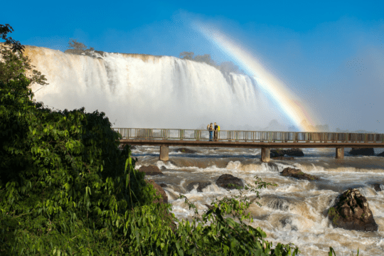 Cataratas do Iguaçu com passarela de visitação, forte volume de água e arco-íris ao fundo