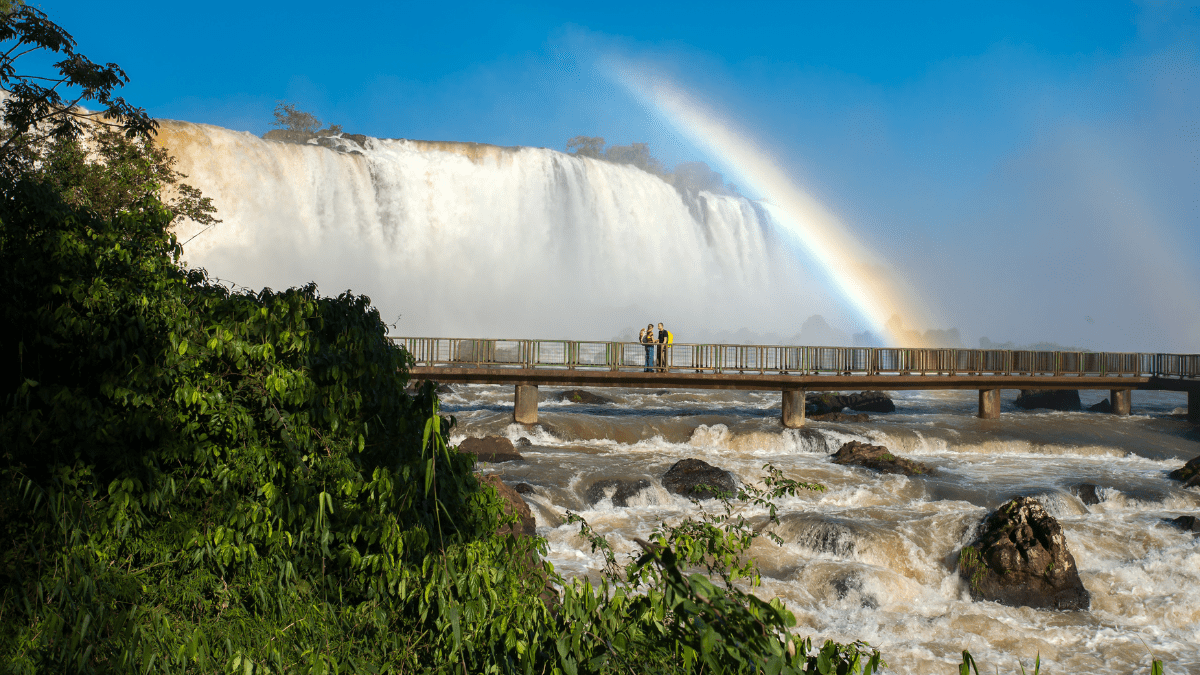 Cataratas do Iguaçu com passarela de visitação, forte volume de água e arco-íris ao fundo